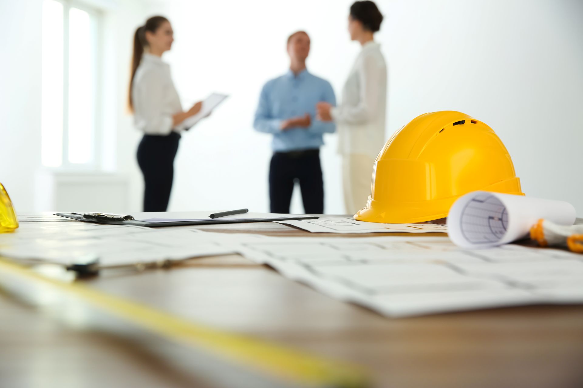 Gelber Schutzhelm auf einem Tisch mit Bauplaenen und Dokumenten, im Hintergrund stehen drei Personen im Gespraech ueber Arbeitsschutz und Sicherheit