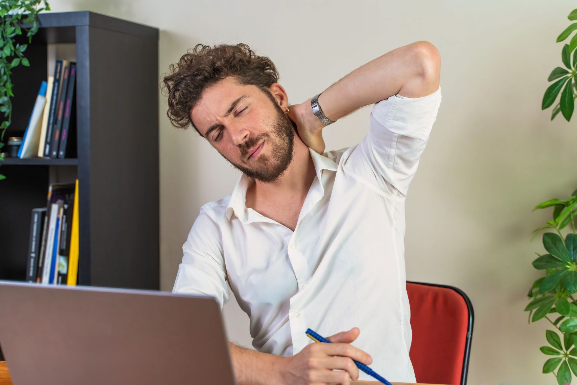 Mann im Buero sitzt am Laptop und haelt sich den Nacken wegen Schmerzen, Beispiel fuer ergonomische Probleme und Arbeitsschutz im Bueroalltag